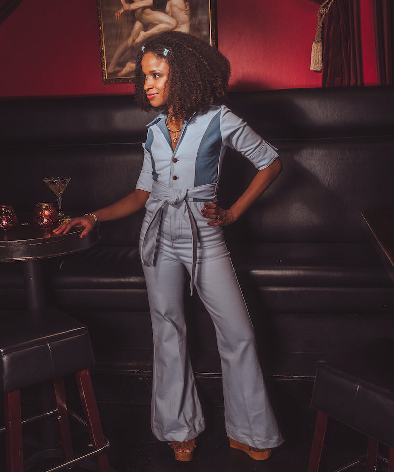Woman in a gray jumpsuit standing in a bar with a painting on the wall.