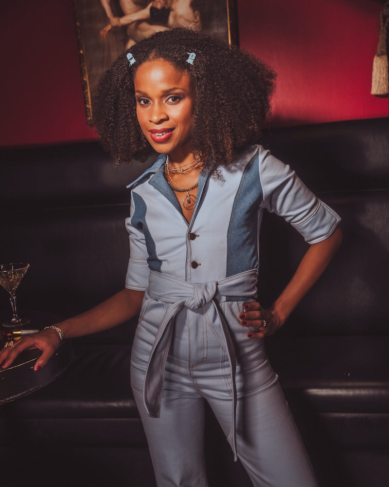 Woman in a gray jumpsuit standing in a bar setting with a drink tray.