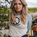 Woman wearing a t-shirt with a logo and text, standing outdoors with greenery and a motorcycle in the background Woman wearing a peace symbol shirt with text outdoors wearing Soul Sister since 1969.