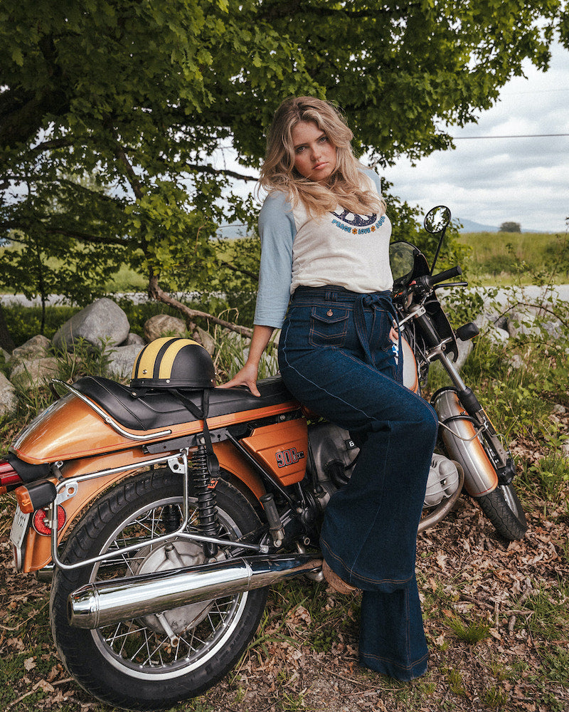 Woman sitting on a motorcycle in a natural setting with trees and grass wearing Soul Sister since 1969.