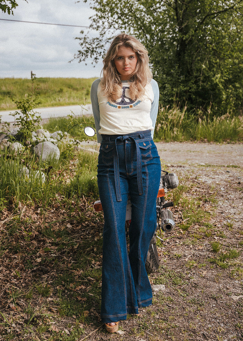 Woman in a white t-shirt and blue jeans standing next to a motorcycle in a natural setting Woman wearing a peace symbol shirt with text outdoors wearing Soul Sister since 1969.