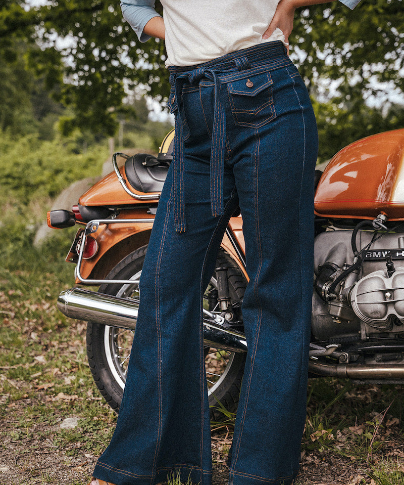 Person wearing blue jeans standing next to an orange motorcycle in a natural setting wearing Soul Sister since 1969.