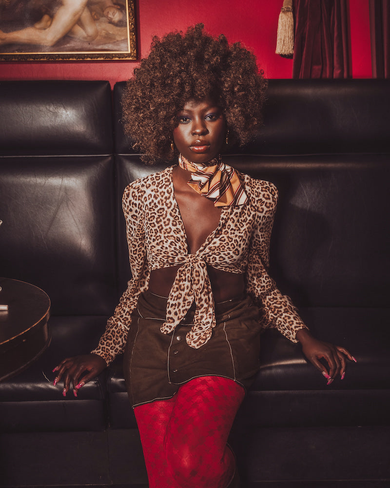Woman in leopard print top and red pants sitting on a black leather couch with a red wall and painting in the background.