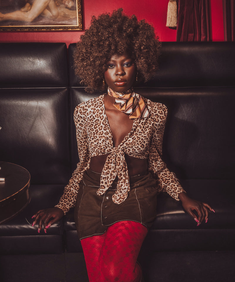 Woman in leopard print top and red pants sitting on a black leather couch with a red wall and painting in the background.