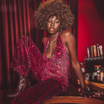 Woman in a red sequin outfit sitting at a bar with red curtains and bottles in the background
