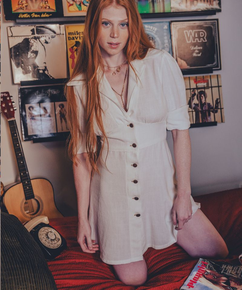 Woman in a white dress standing in a room with vinyl records and a guitar.
