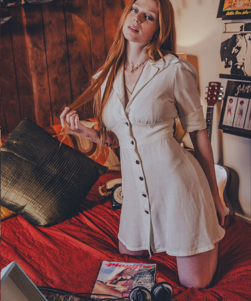 Woman in a white dress sitting on a bed in a room with wooden walls and posters.