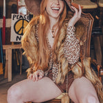 Woman with long red hair wearing a wide-brimmed hat and leopard print top, sitting on a wooden chair.