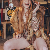 Woman with long red hair wearing a wide-brimmed hat and leopard print top, sitting on a wooden chair.