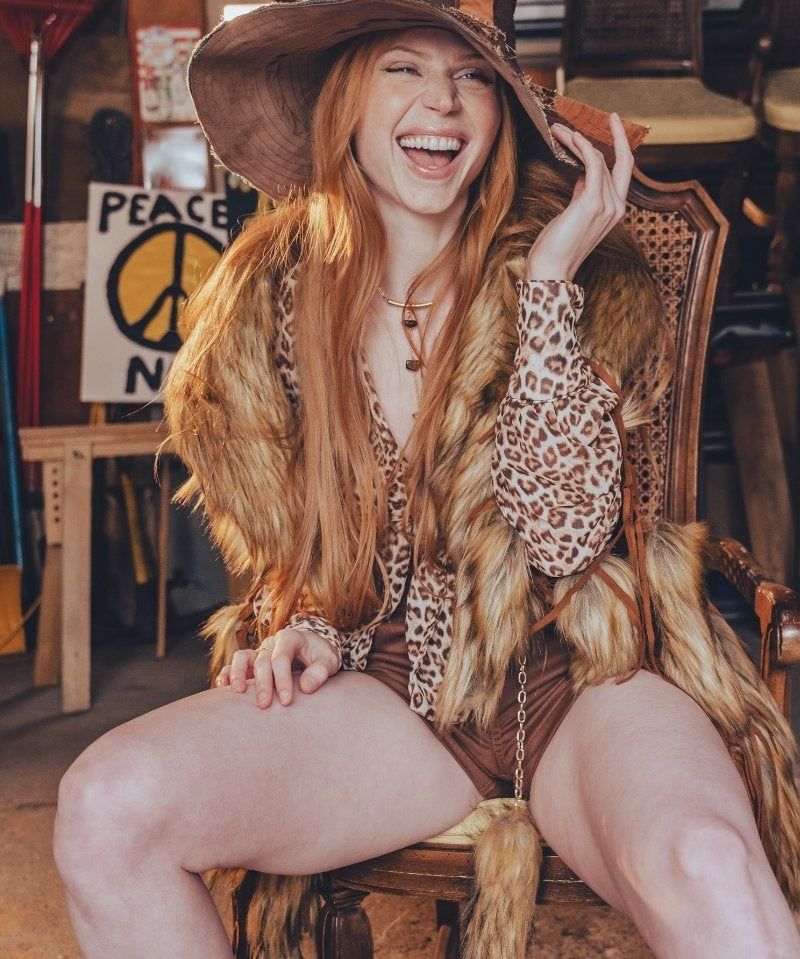 Woman with long red hair wearing a wide-brimmed hat and leopard print top, sitting on a wooden chair.