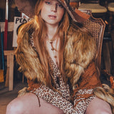 Person wearing a fur-trimmed outfit with a hat, sitting in a rustic setting.