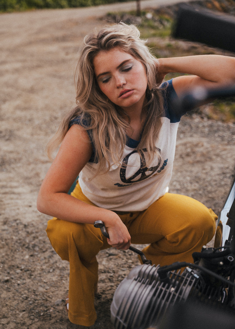 Woman in a sleeveless top and yellow pants sitting on a motorcycle outdoors wearing Soul Sister since 1969.