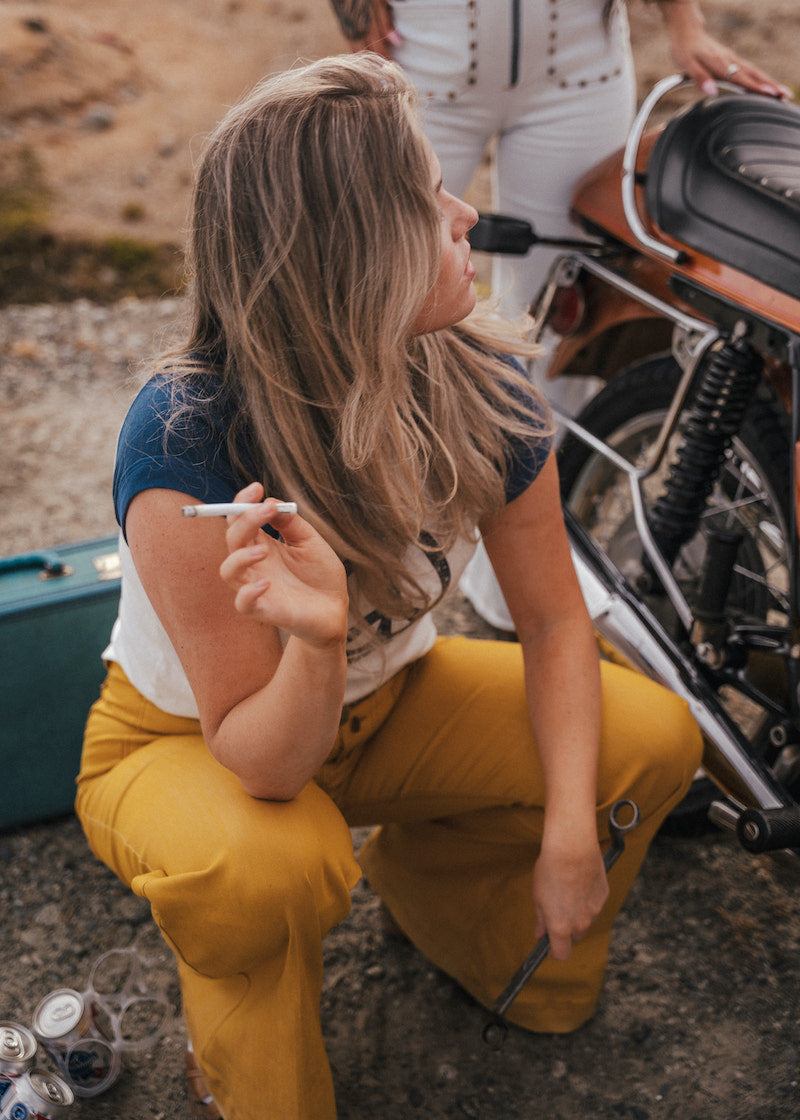Woman in yellow pants sitting next to a motorcycle with a cigarette in her hand wearing Soul Sister since 1969.