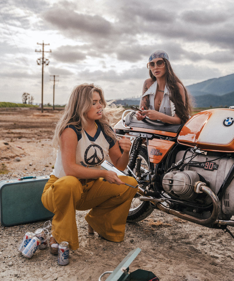 Two women sitting next to a motorcycle in an open area with mountains in the background wearing Soul Sister since 1969.