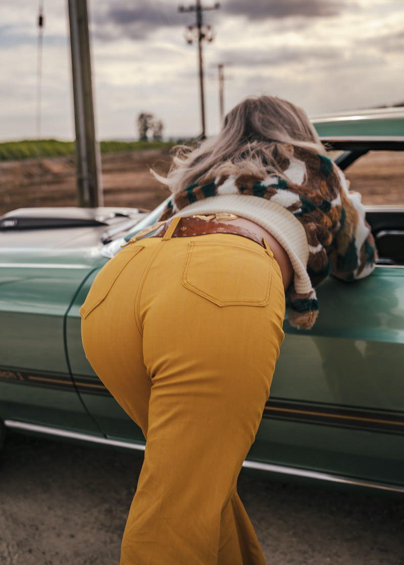Person in yellow pants with a cow on their back, standing next to a green car in an open field wearing soul sister since 1969.