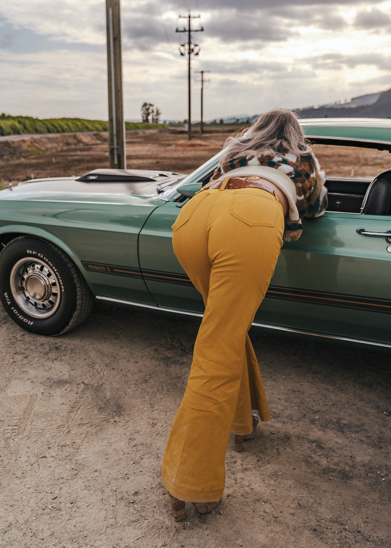 Person wearing mustard yellow flared pants standing next to a green car in an open field wearing soul sister since 1969.