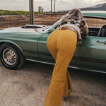 Person wearing mustard yellow flared pants standing next to a green car in an open field wearing soul sister since 1969.
