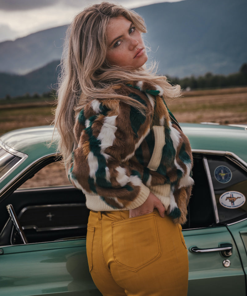 Woman in a patterned jacket and yellow pants standing next to a vintage car with mountains in the background wearing Soul Sister since 1969