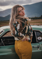 Woman in a patterned jacket and yellow pants standing next to a vintage car with mountains in the background wearing Soul Sister since 1969