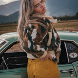 Woman in a patterned jacket and yellow pants standing next to a vintage car with mountains in the background v
