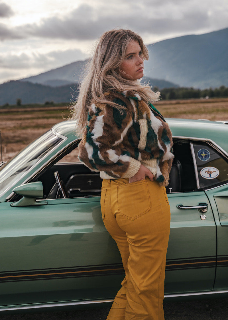 Woman in a patterned jacket and yellow pants standing next to a vintage car with mountains in the background wearing Soul Sister since 1969