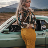 Woman in a patterned jacket and yellow pants standing next to a vintage car with mountains in the background v