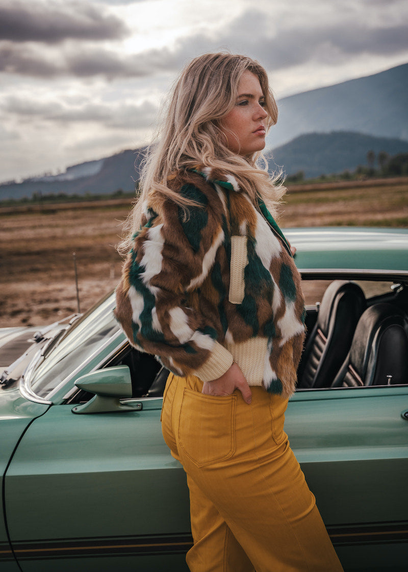 Woman in a patterned coat and yellow pants standing next to a vintage car with mountains in the background wearing Soul Sister since 1969