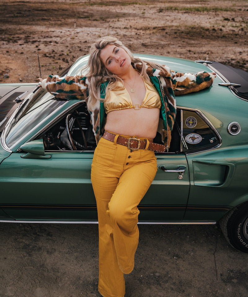 Woman in a gold top and yellow pants standing next to a vintage car in an open field wearing soul sister since 1969.