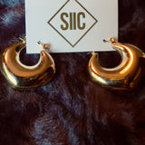 Gold hoop earrings on a dark fur surface with a card displaying 'SIIIC'.