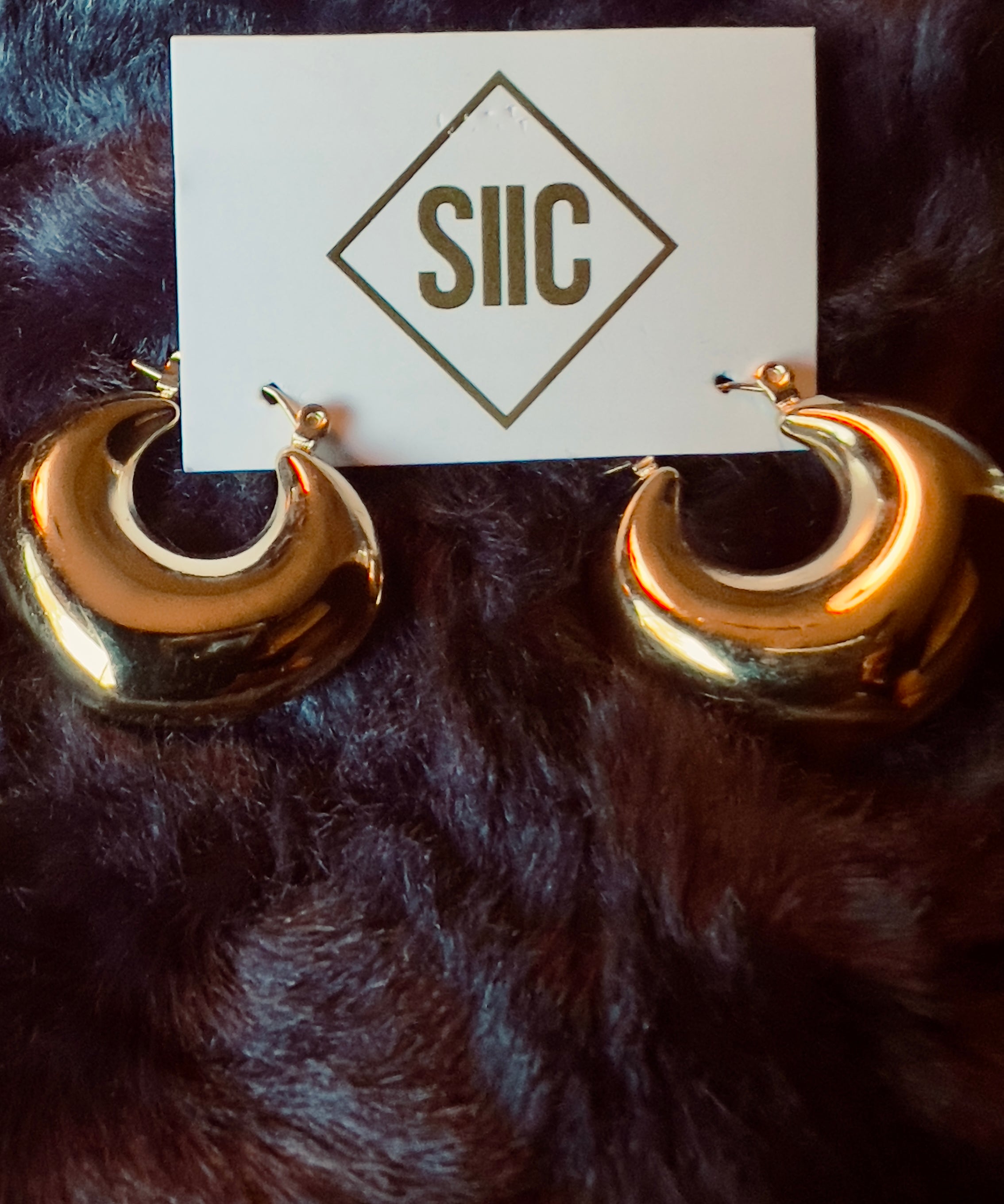 Gold hoop earrings on a dark fur surface with a card displaying 'SIIIC'.