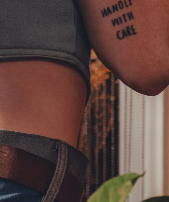 Person wearing a tattoo on their arm with the text 'HANDLE WITH CARE' against a blurred background.