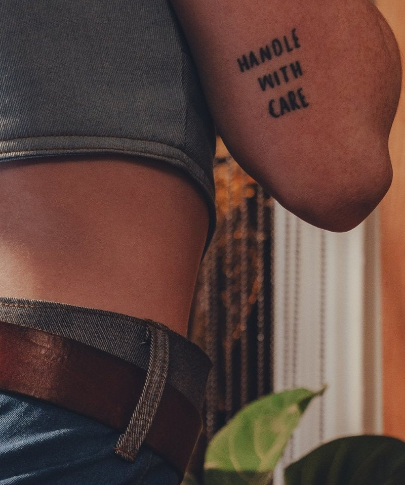Person wearing a tattoo on their arm with the text 'HANDLE WITH CARE' against a blurred background.