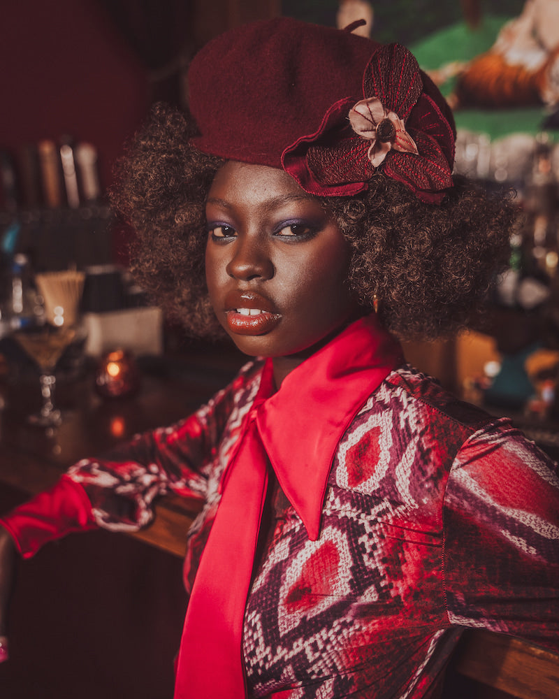 Woman wearing a red hat with a flower and patterned outfit in a bar setting
