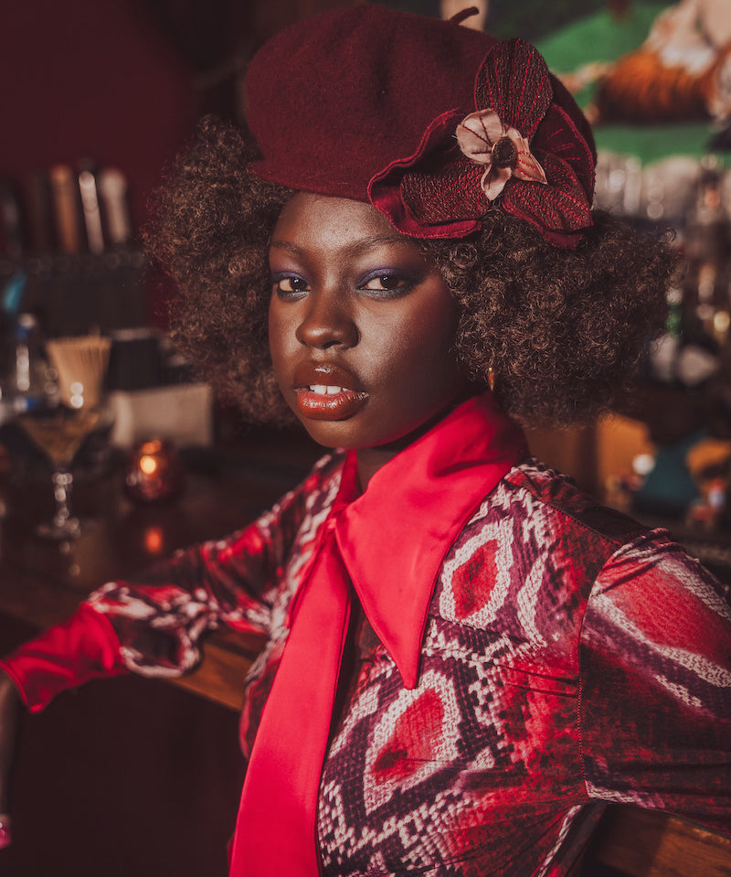 Woman wearing a red hat with a flower and patterned outfit in a bar setting