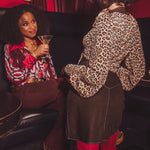 Two women in a social setting, one holding a cocktail glass, in a dimly lit room.