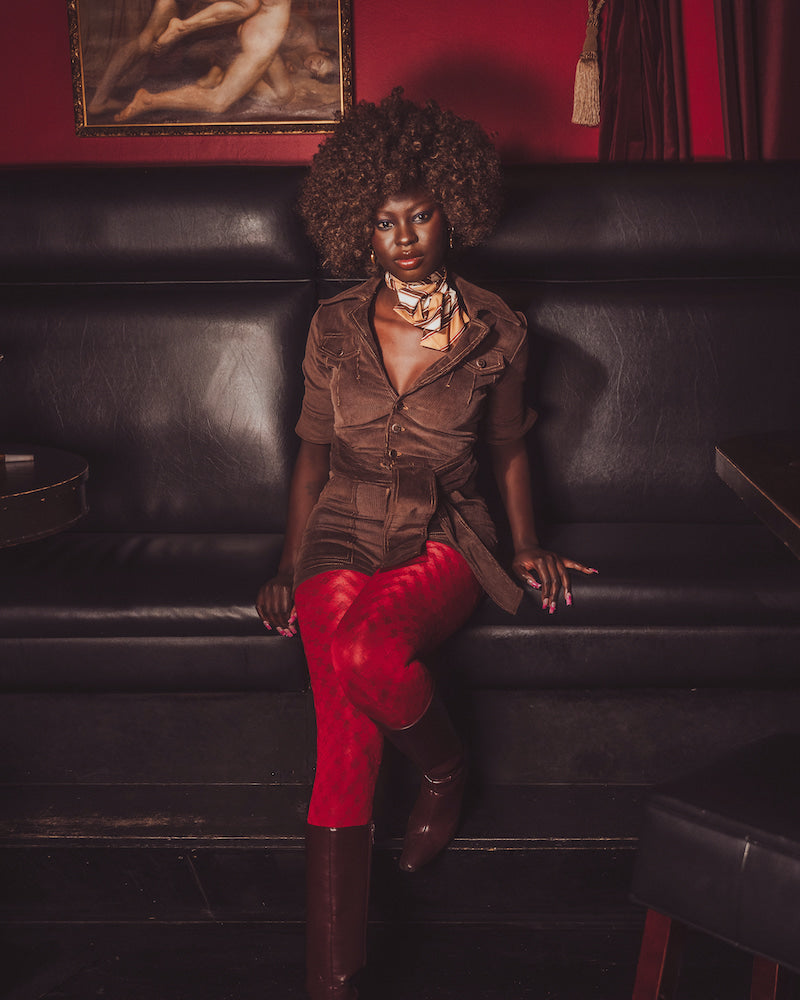 Woman sitting on a black leather couch wearing a brown jacket and red pants, with a red wall and painting in the background.