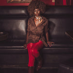 Woman sitting on a black leather couch wearing a brown jacket and red pants, with a red wall and painting in the background.