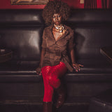 Woman sitting on a black leather couch wearing a brown jacket and red pants, with a red wall and painting in the background.