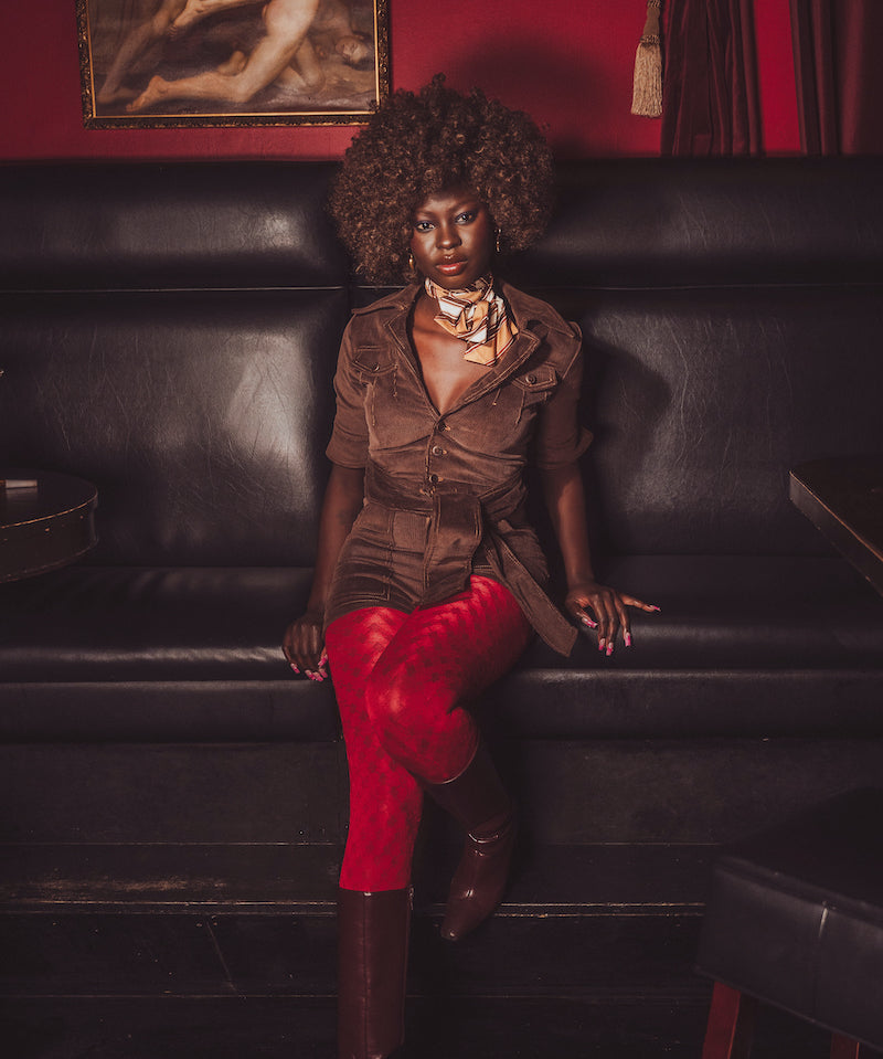 Woman sitting on a black leather couch wearing a brown jacket and red pants, with a red wall and painting in the background.