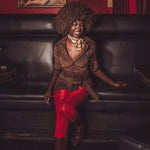 Woman in a brown jacket and red pants sitting on a black leather couch with a red wall and painting in the background.