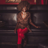 Woman in a brown jacket and red pants sitting on a black leather couch with a red wall and painting in the background.
