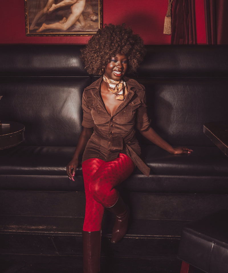 Woman in a brown jacket and red pants sitting on a black leather couch with a red wall and painting in the background.