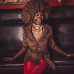 Woman in a brown jacket and red pants sitting on a black leather couch with a painting in the background.