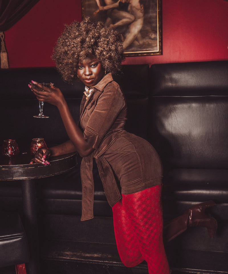 Woman in a brown top and red pants holding a glass in a dimly lit room with a painting on the wall.