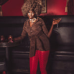 Woman in a brown coat and red pants holding a cocktail in a dimly lit room.