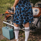 Person wearing a blue patterned dress standing next to a vintage motorcycle with a suitcase nearby wearing Soul Sister since 1969..