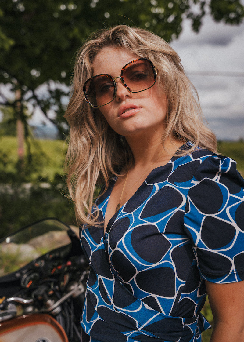 Woman wearing sunglasses and a blue patterned dress standing outdoors with greenery in the background wearing Soul Sister since 1969.