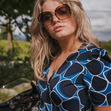 Woman wearing sunglasses and a blue patterned dress standing outdoors with greenery in the background wearing Soul Sister since 1969.