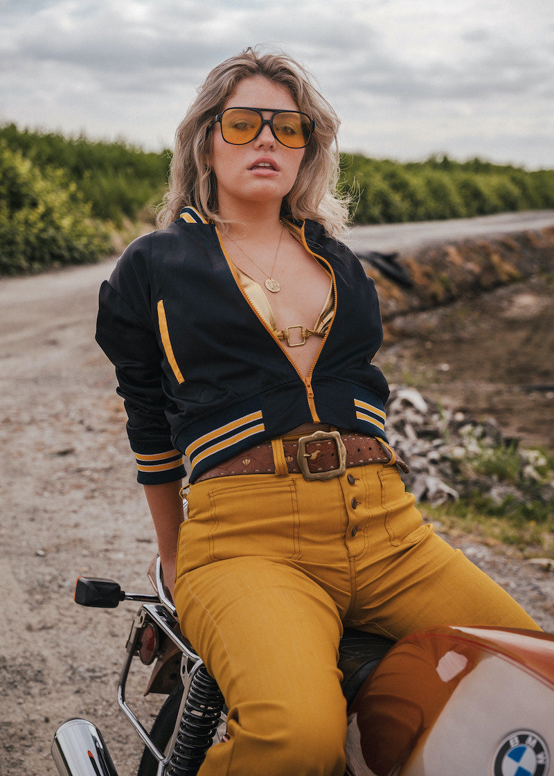 Woman in a black and yellow jacket and mustard pants sitting on a motorcycle in a rural setting Soul Sister since 1969 .