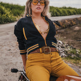 Woman in a black and yellow jacket and mustard pants sitting on a motorcycle in a rural setting Soul Sister since 1969 .